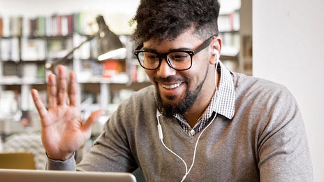 Smiling man waving at his laptop while attending Italian classes online with headphones on in a cozy library café setting.