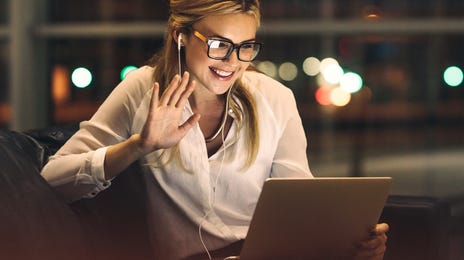 Woman taking online language lessons via video call at night, smiling and waving at her laptop with earphones in.